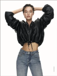 Model wearing a black leather jacket and blue denim jeans posing against a white background with hands behind head
