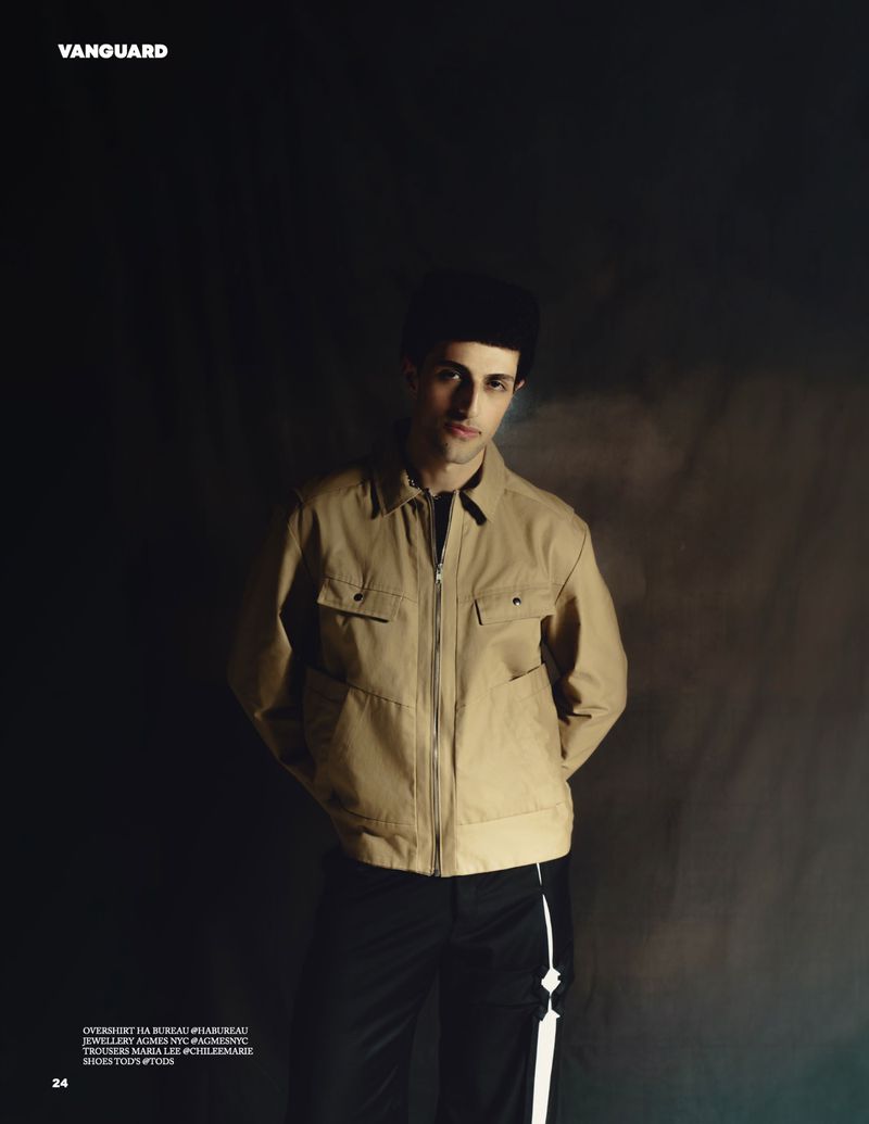 Man wearing a camel jacket and black pants against a dark textured background for Vanguard Magazine.