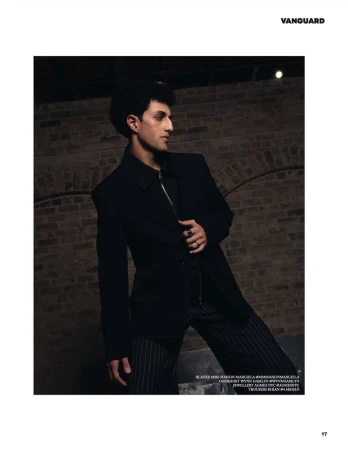 A man in a black jacket and pinstripe trousers posing against a brick wall for Vanguard Magazine.