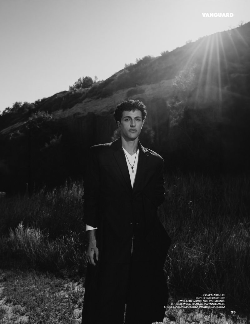 Monochrome editorial fashion portrait of a man in a dark blazer standing outdoors against a grassy slope under bright sunlight.