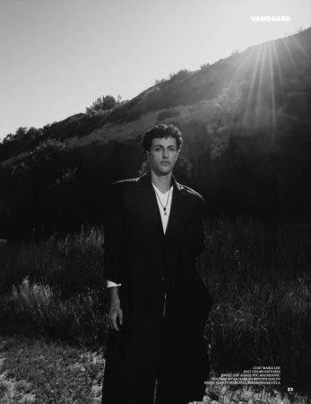 Monochrome editorial fashion portrait of a man in a dark blazer standing outdoors against a grassy slope under bright sunlight.