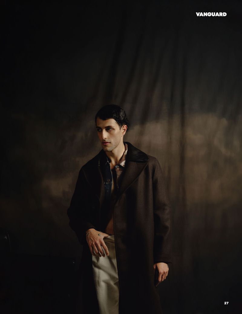 Man wearing a dark coat and light trousers standing against a textured dark backdrop in an editorial portrait.