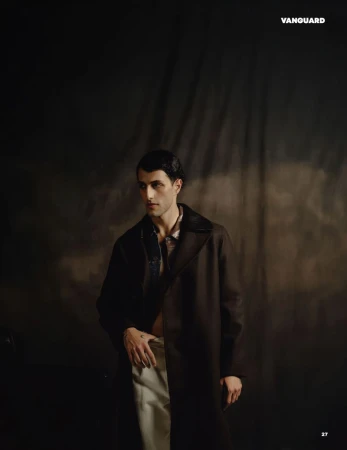 Man wearing a dark coat and light trousers standing against a textured dark backdrop in an editorial portrait.