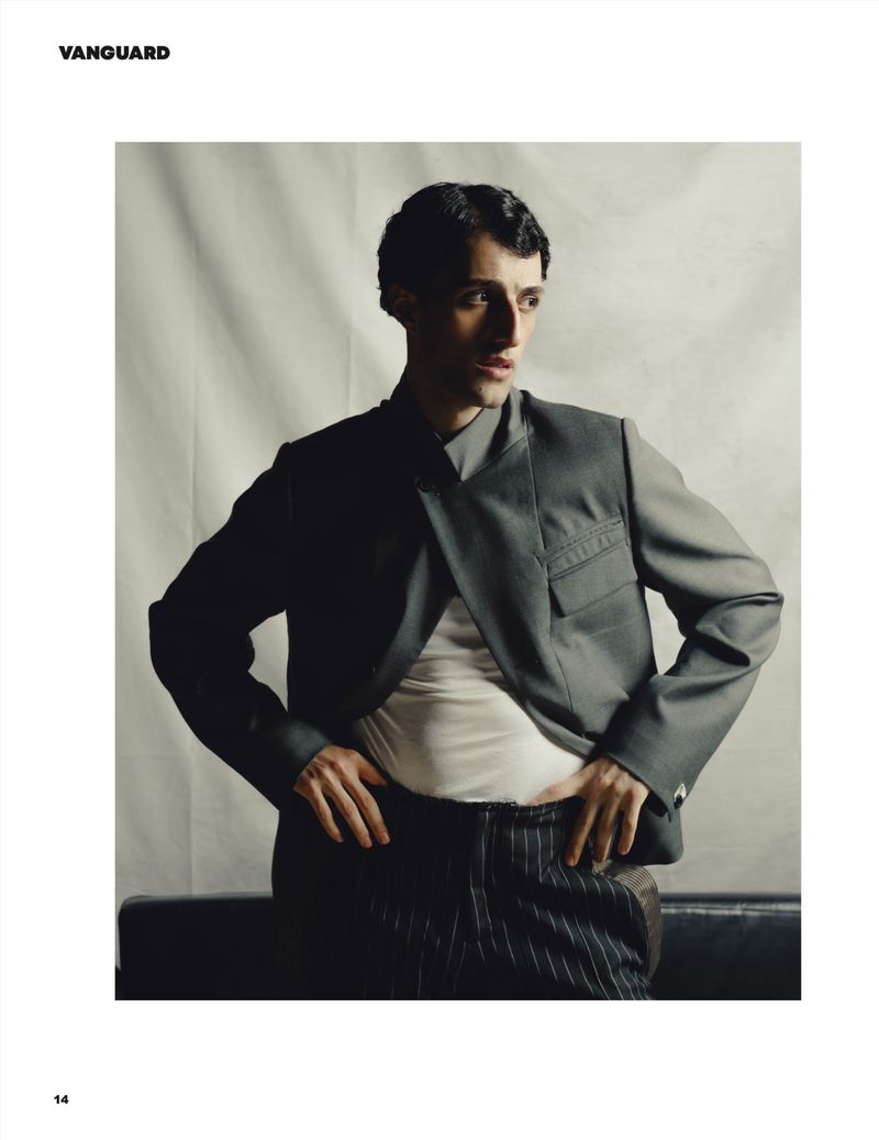 Man wearing a grey structured jacket and pinstripe trousers posing against a textured backdrop for Vanguard Magazine.