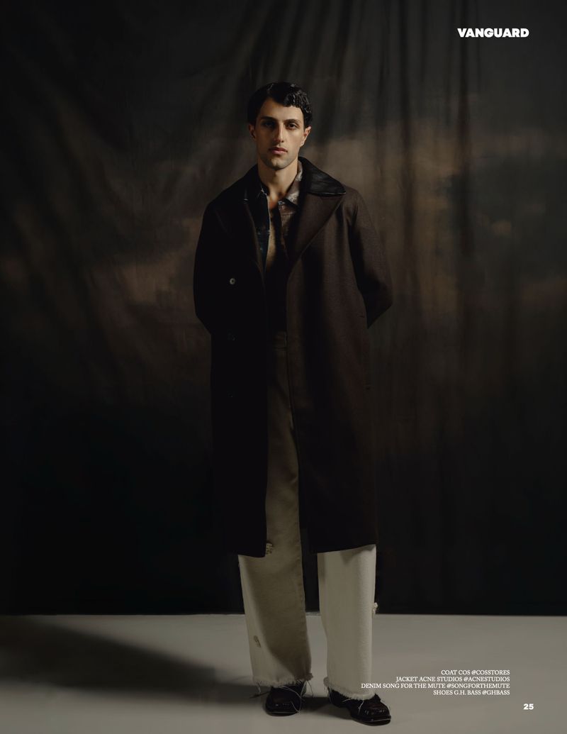 Man wearing a dark overcoat and light trousers standing against a textured backdrop for Vanguard Magazine