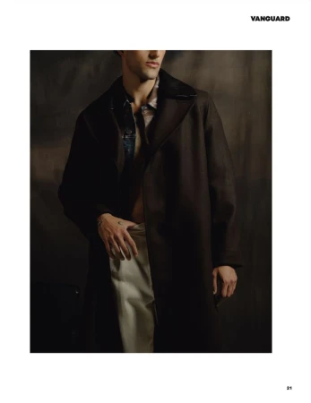 Man wearing a dark brown coat over light trousers against a textured dark backdrop for Vanguard Magazine