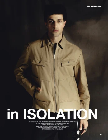 Man wearing a camel zip jacket and dark trousers standing against a textured dark background for Vanguard Magazine.