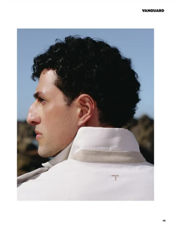 Profile portrait of a man with dark curly hair wearing a white high-collar jacket against a clear sky and rocky background