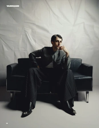 Man seated on a black leather sofa wearing a dark jacket and wide pinstripe trousers against a rumpled white backdrop