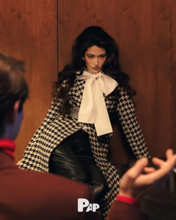 Woman in a black and white houndstooth coat with a large white bow tie seated against a dark wood wall