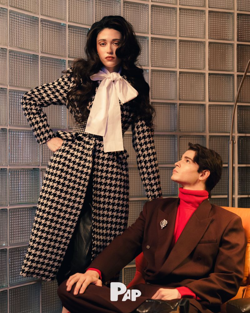 Woman in houndstooth coat with large white bow standing next to a seated man in a brown blazer and red turtleneck for PAP Magazine.