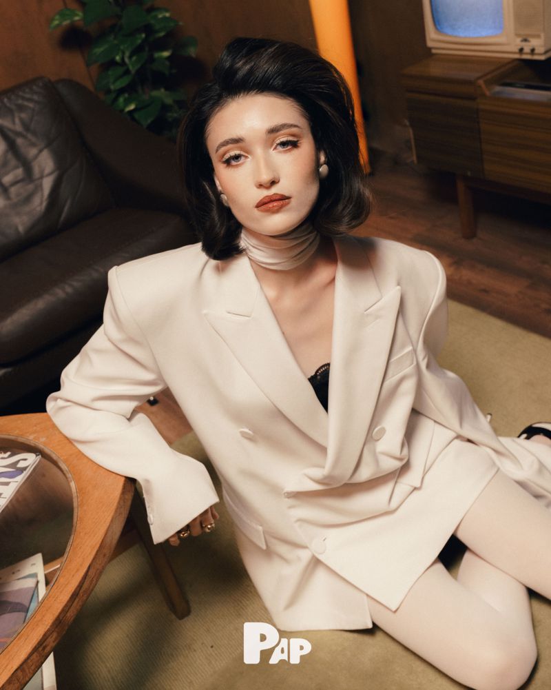Woman in white double breasted blazer and white tights seated on carpet in a retro styled interior living room.