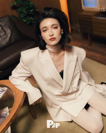 Woman in white double breasted blazer and white tights seated on carpet in a retro styled interior living room.