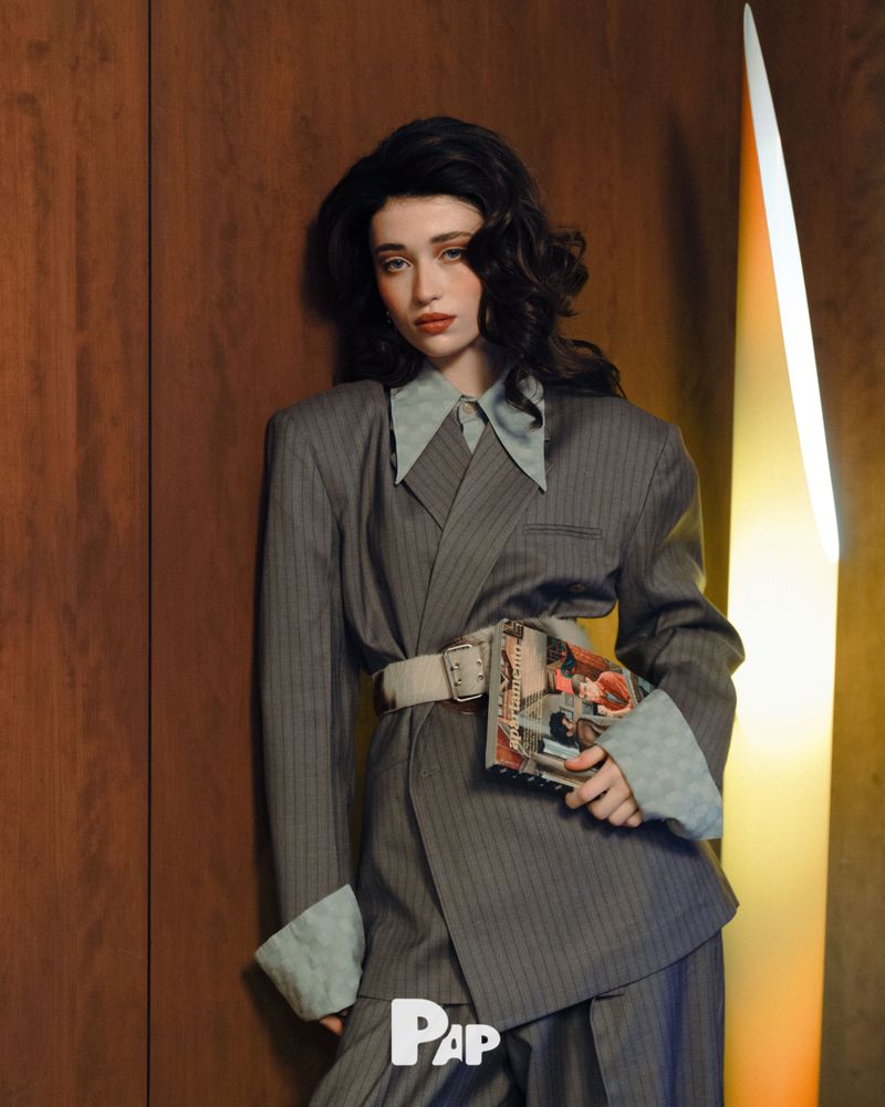 A woman in a grey pinstripe suit and wide belt holding a magazine against a wooden wall with a vertical light fixture
