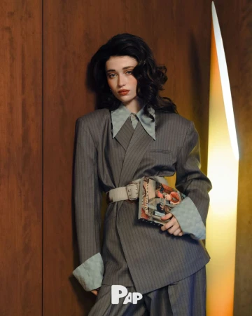 A woman in a grey pinstripe suit and wide belt holding a magazine against a wooden wall with a vertical light fixture