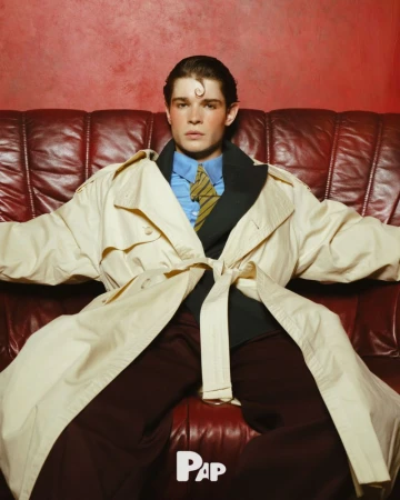 Young man in a beige trench coat and blue shirt seated on a deep red leather sofa for PAP Magazine