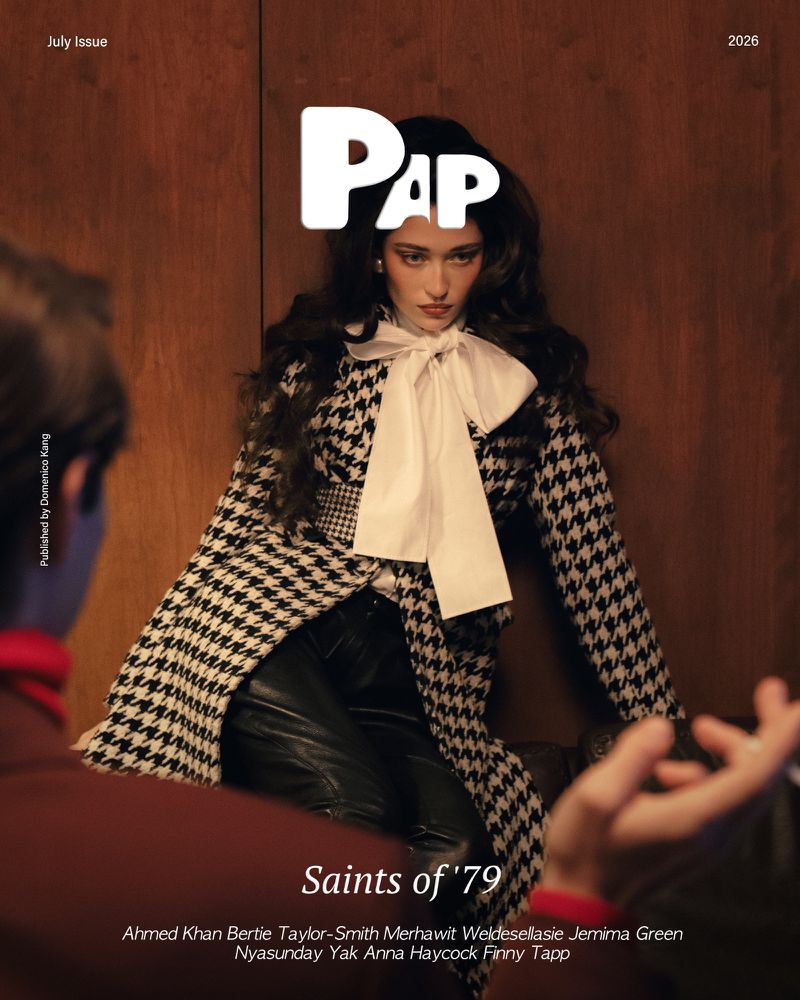 A woman in a houndstooth coat with a large white bow tie seated against a wood-paneled wall, with a man in the foreground.