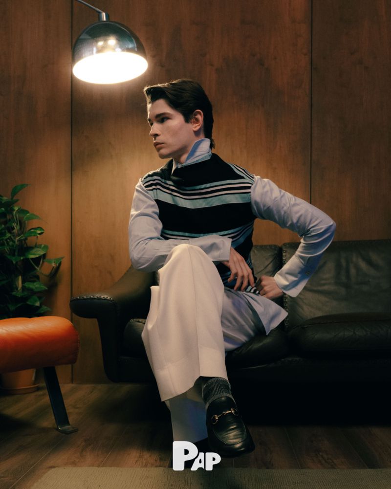 Young man seated on a dark leather sofa wearing a striped vest over a light blue shirt and white trousers, under a warm overhead lamp.
