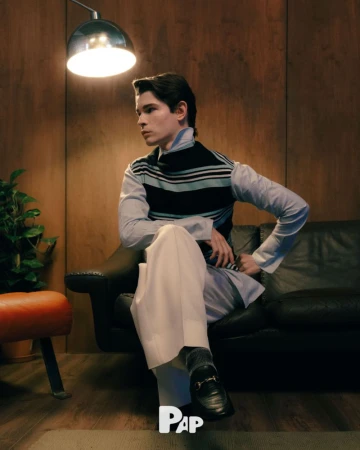 Young man seated on a dark leather sofa wearing a striped vest over a light blue shirt and white trousers, under a warm overhead lamp.
