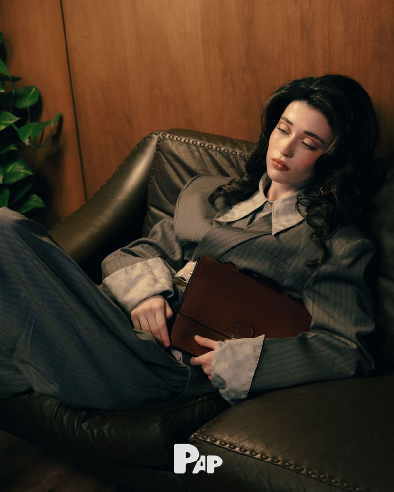 Woman in pinstripe blazer holding a brown leather briefcase while lounging on a dark leather sofa for PAP Magazine