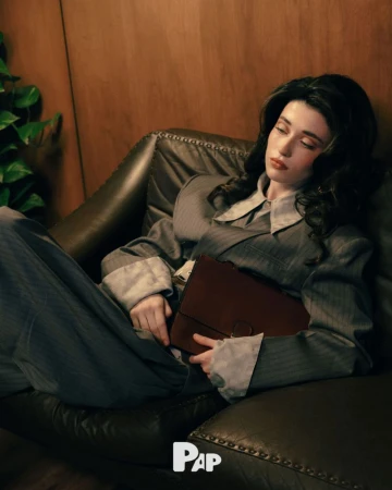 Woman in pinstripe blazer holding a brown leather briefcase while lounging on a dark leather sofa for PAP Magazine