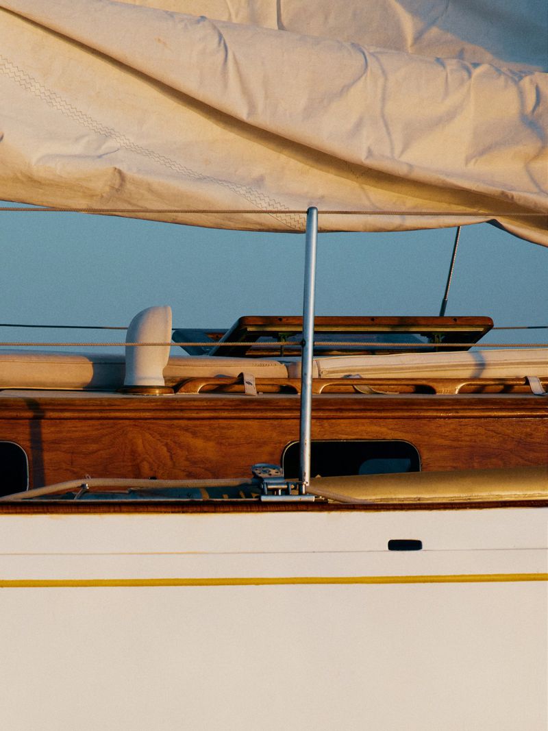 Close-up of a sailboat with a white sail, wooden deck, and polished metal hardware, bathed in warm light against a clear blue sky.