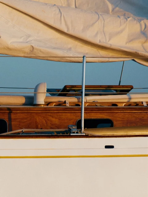 Close-up of a sailboat with a white sail, wooden deck, and polished metal hardware, bathed in warm light against a clear blue sky.
