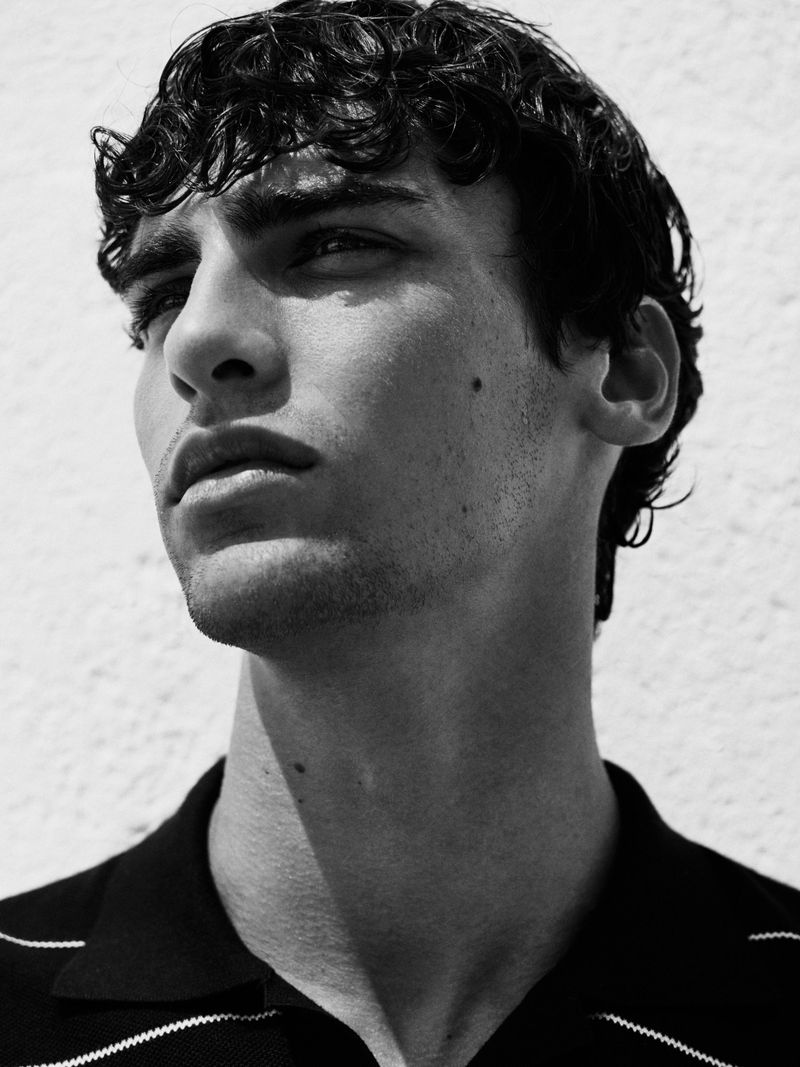 Portrait of a young adult man with curly dark hair, wearing a dark and white striped polo shirt, against a white wall background.