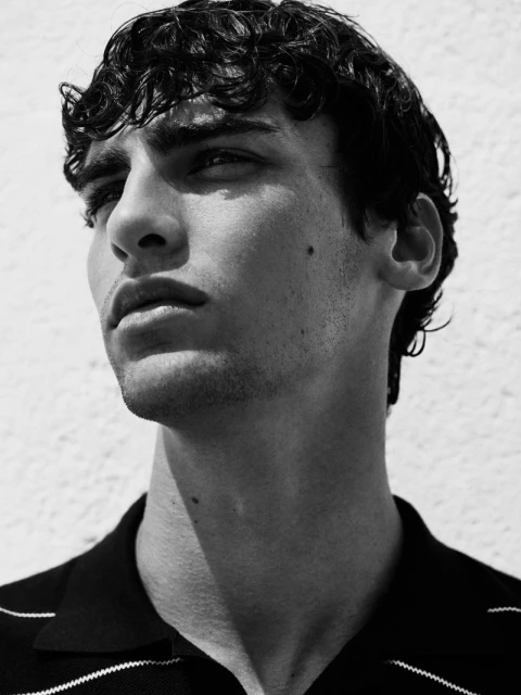 Portrait of a young adult man with curly dark hair, wearing a dark and white striped polo shirt, against a white wall background.