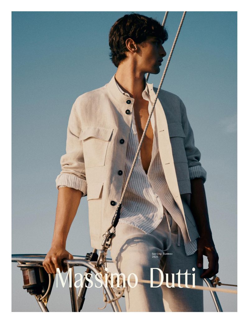 A tanned male fashion model poses on a sailboat, wearing a cream-colored linen jacket and pants with an unbuttoned striped shirt and bare chest.