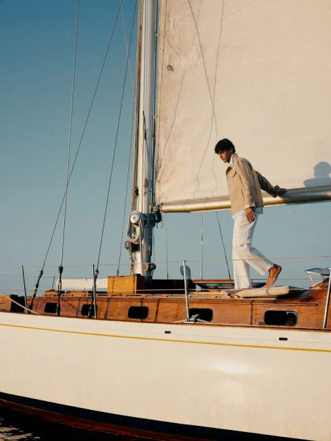 A stylish man in a tan jacket and white pants stands on the side of a white sailboat with a large sail against a clear blue sky.