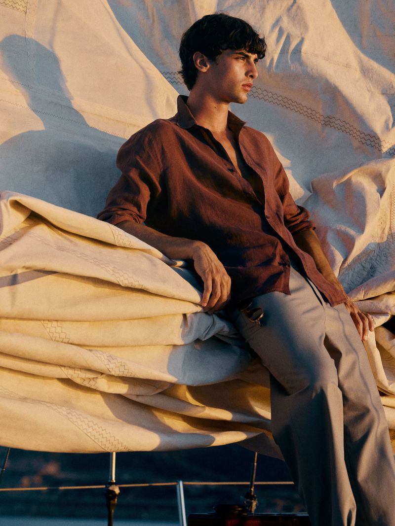 A young man sits on a boat draped with sails in a brown button-down shirt and grey slacks while illuminated by warm golden sunlight.