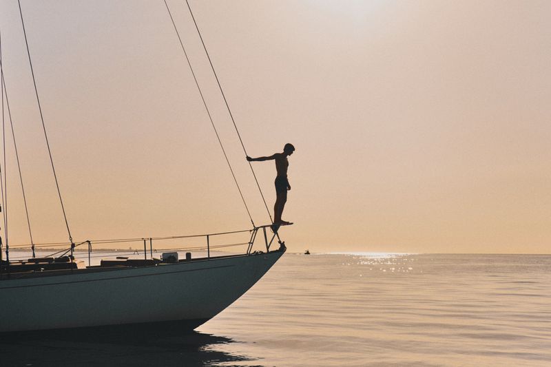 Silhouette of shirtless male standing on the bow of a sailboat at sea, facing the ocean at sunset. Boat, horizon, water.