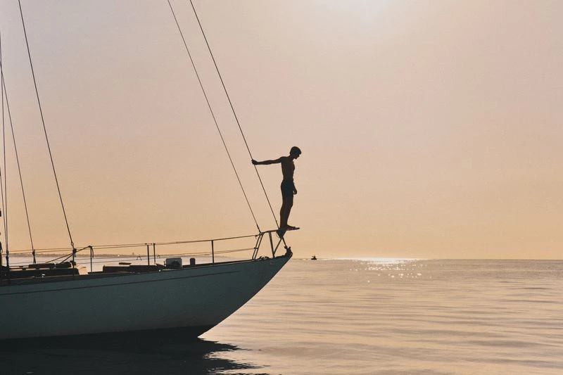 Silhouette of shirtless male standing on the bow of a sailboat at sea, facing the ocean at sunset. Boat, horizon, water.
