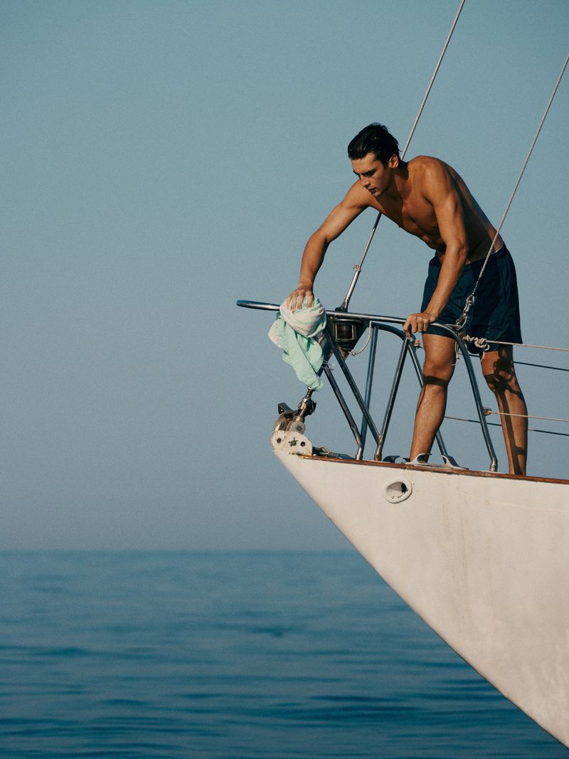 Shirtless man on boat cleaning railing with cloth in bright daylight over blue sea; nautical lifestyle, travel and transportation.
