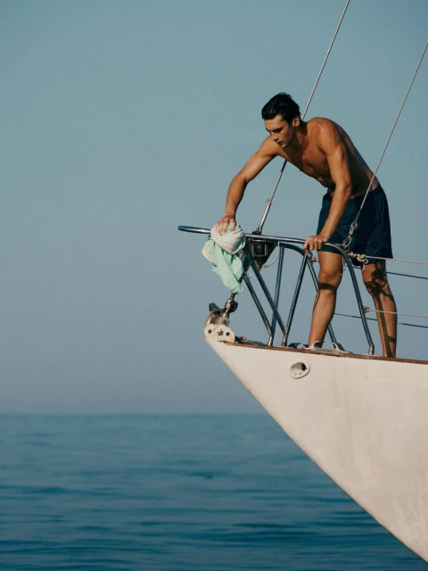 Shirtless man on boat cleaning railing with cloth in bright daylight over blue sea; nautical lifestyle, travel and transportation.