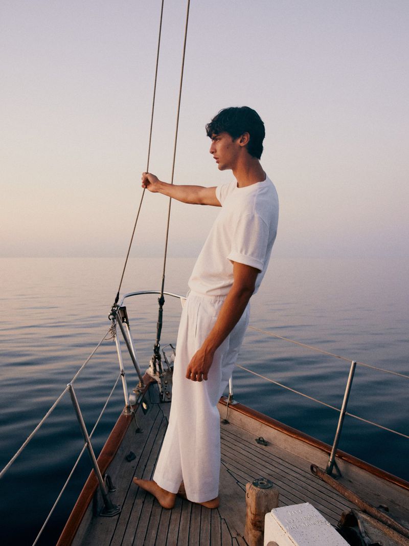 A young adult man stands on a sailboat wearing a white linen outfit, looking towards the horizon, with the ocean and sky in the background.