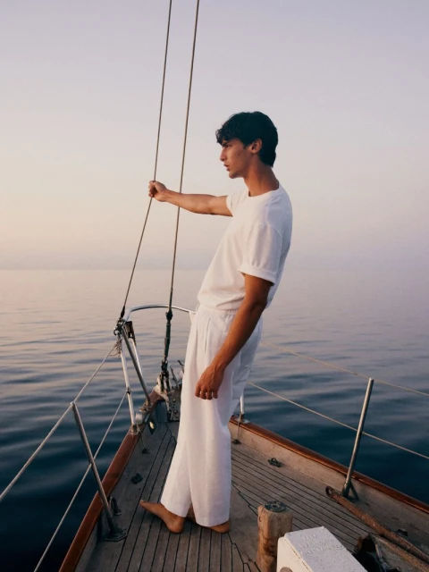 A young adult man stands on a sailboat wearing a white linen outfit, looking towards the horizon, with the ocean and sky in the background.