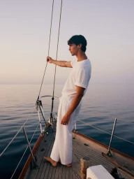 Man wearing white t-shirt and white trousers standing on sailboat deck at sunset