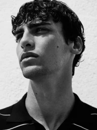 Black and white portrait of a man looking upward against a textured white wall wearing a dark collared polo shirt.