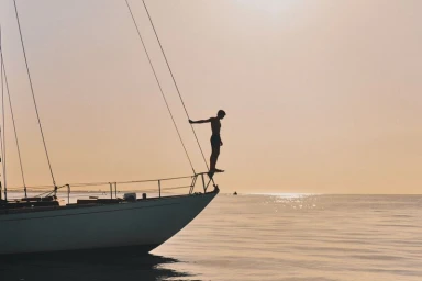 Silhouette of a man standing on the bow of a sailboat over calm ocean water at sunset.