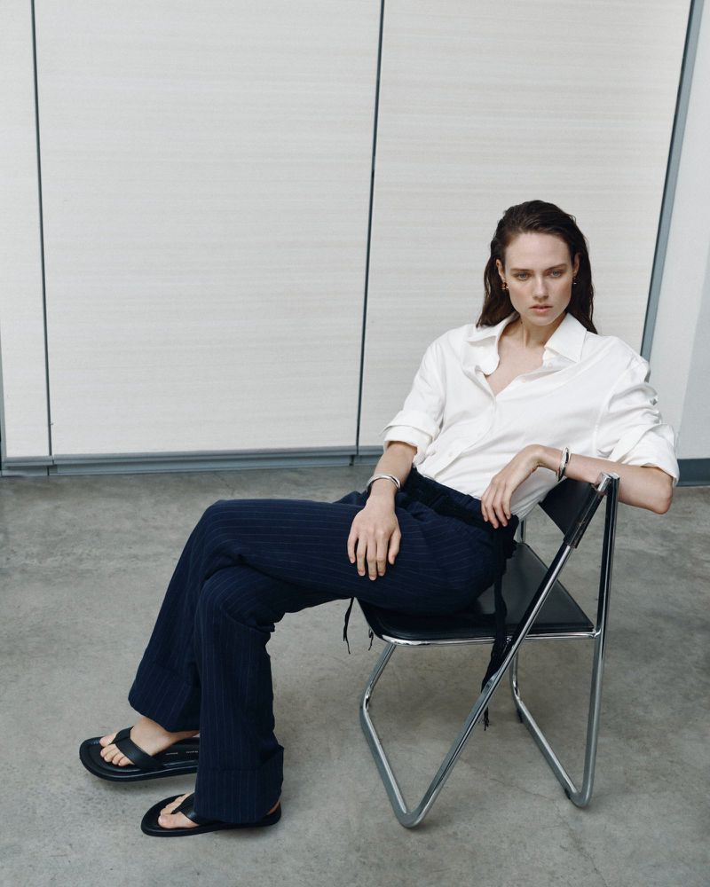 Model Kate McNamara seated on a black chair wearing a white button-down shirt and dark trousers for ACCORDA SS27