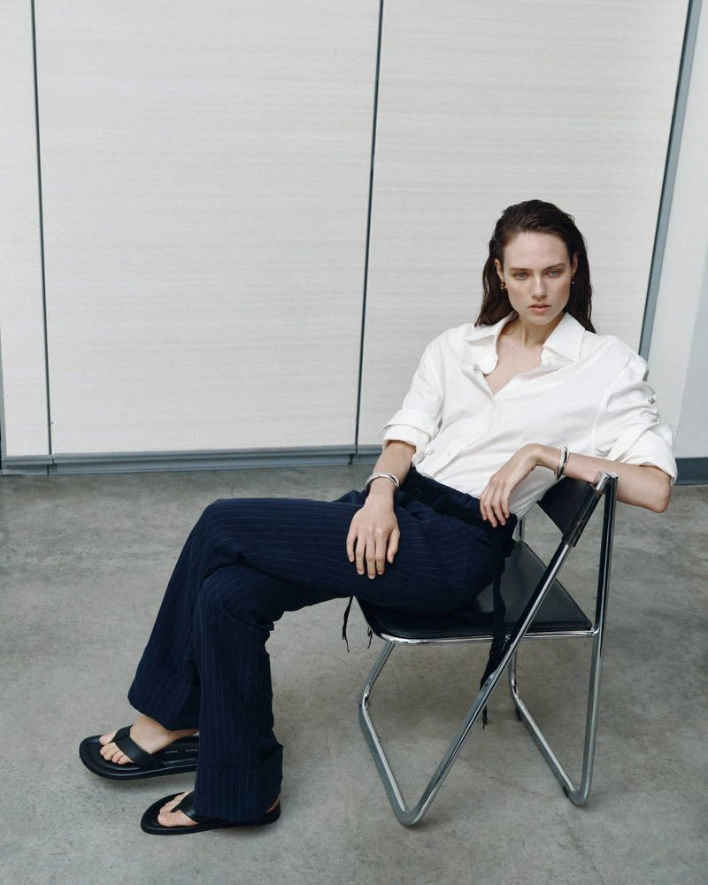 Model Kate McNamara seated on a black chair wearing a white button-down shirt and dark trousers for ACCORDA SS27