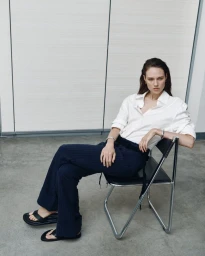 Model Kate McNamara seated on a black chair wearing a white button-down shirt and dark trousers for ACCORDA SS27