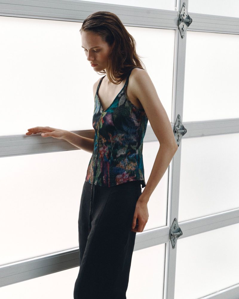 Model Kate McNamara wearing a dark patterned tank top and black trousers against a large white frosted window.