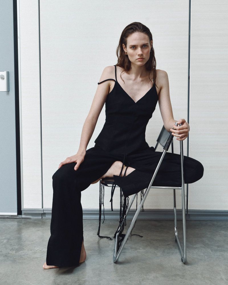 Model Kate McNamara seated on a metal folding chair wearing a black slip dress against a paneled wall.