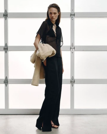 Model in black sheer shirt and trousers holding a beige textured coat against a gridded glass and white background.