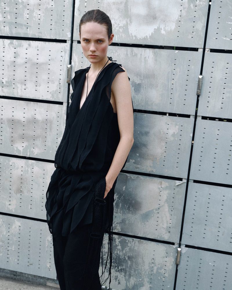 Model wearing a black sleeveless tunic top and matching trousers against an industrial grey panel background.