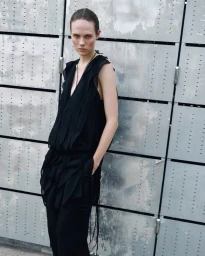 Model wearing a black sleeveless tunic top and matching trousers against an industrial grey panel background.