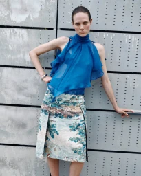Model wearing a blue sleeveless ruffled top and patterned landscape skirt posing against a corrugated metal wall.